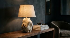 A sculptural table lamp crafted from raw beige travertine stone with a linen shade, resting on a dark walnut console table next to stacked books.